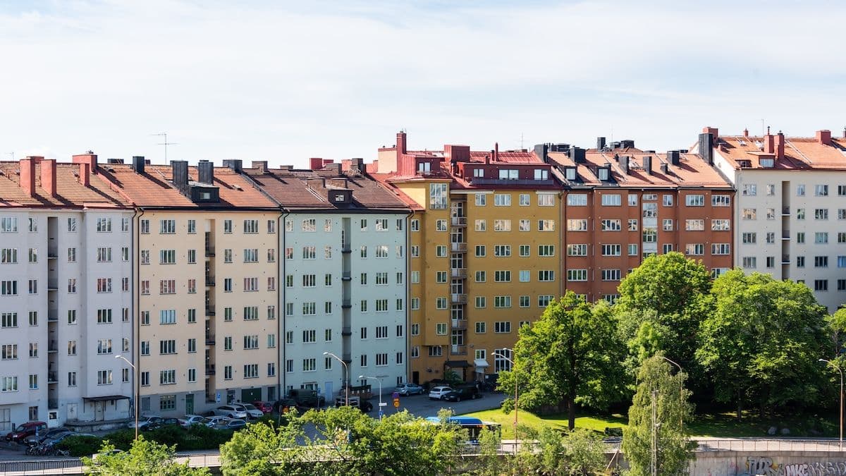 Bild på avstånd av flera, färgglada lägenhetshus en solig dag. Framför husen är grönt trädområde.