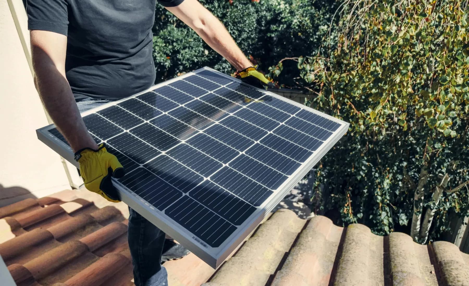 En solcellsinstallatör håller i solpaneler uppe på ett tak i solljus.