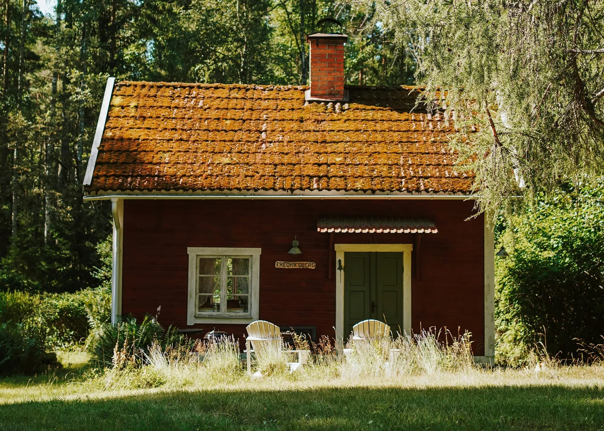 Röd stuga med vita knutar i skogsbrynet med trädgårdsmöbler utanför.