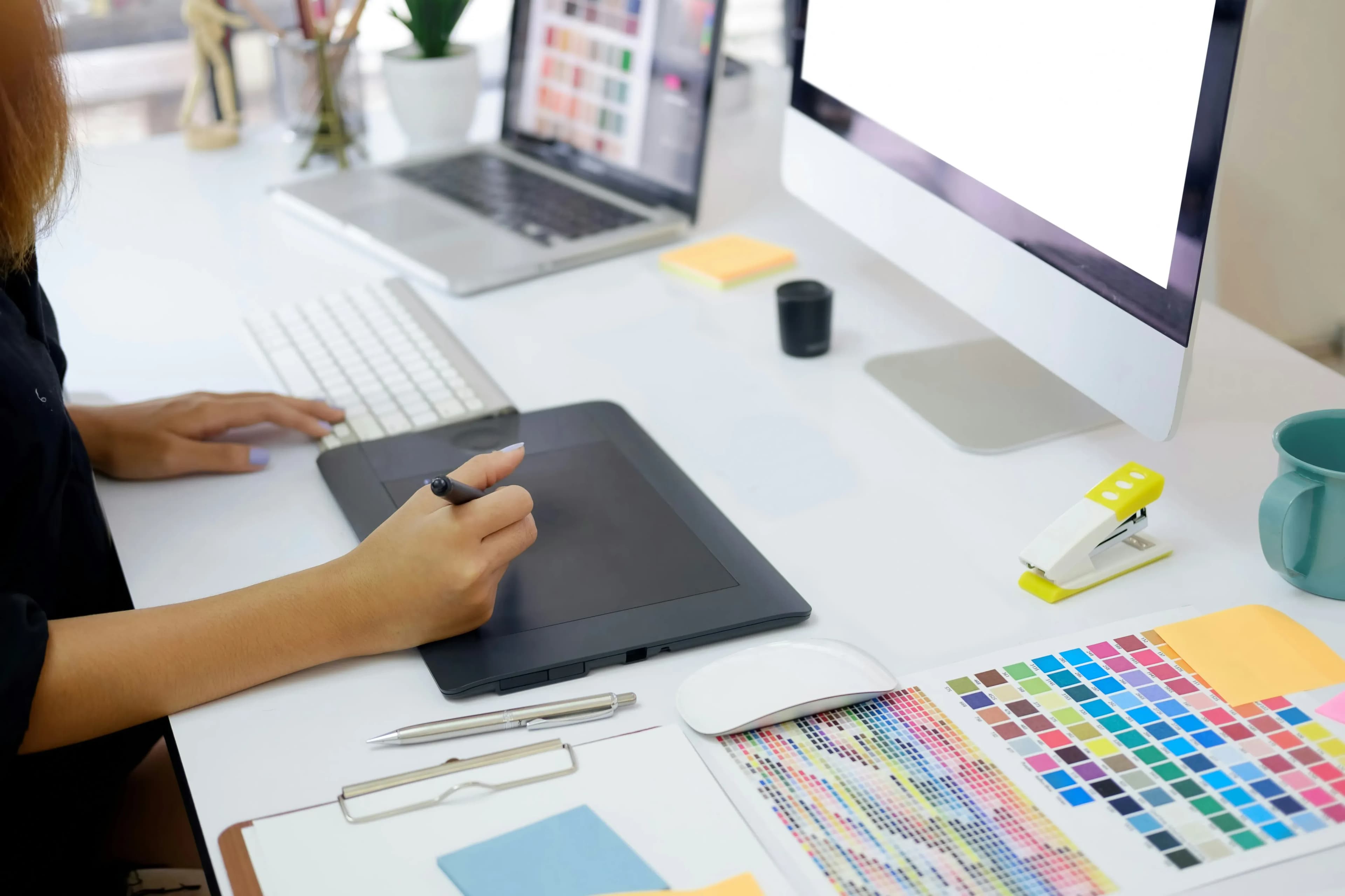 Person using a graphic tablet at a desk with a laptop, computer monitor, color swatches, and office supplies.
