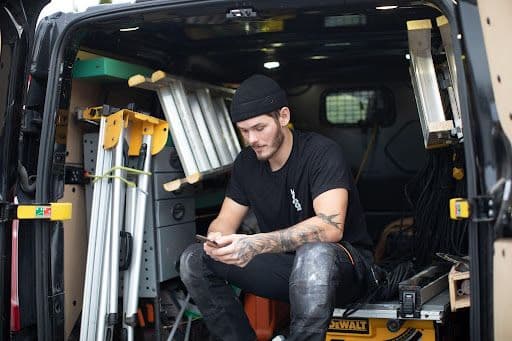Kille som sitter i bakluckan på en byggbil med verktyg och stegar och sitter med mobilen