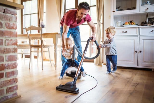 Vuxen man dammsuger med två små barn hemma i köket eller vardagsrummet