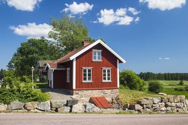 Röd sommarstuga på stenvall med buskar och träd runtomkring