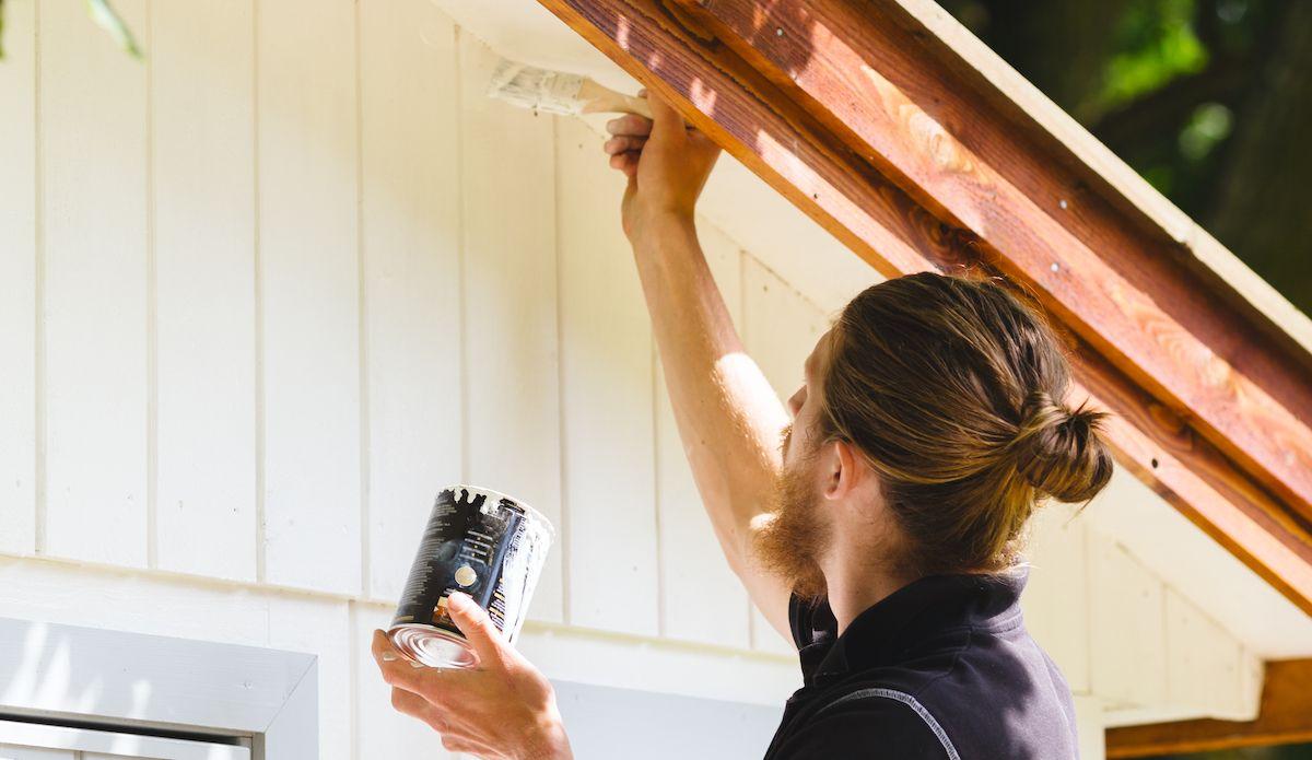 Målare målar ett hus vitt med pensel strax under taknocken som går i träröd ton.