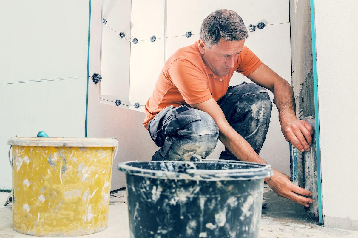 En hantverkare som jobbar med att sätta kakel i ett badrum. Framför honom står stora hinkar med spackel.