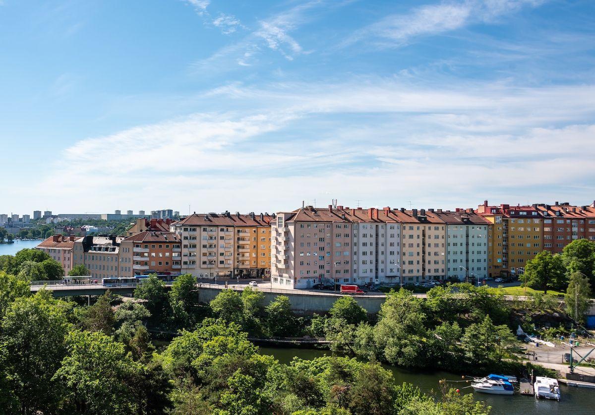 Utsikt över flerfärgade lägenhetshus vid Lilla Essingen i Stockholm. Framför husen är det grönska och ovanför en blå himmel.