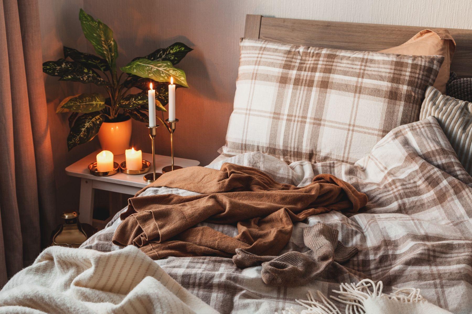 Mysig säng med tända ljus på nattduksbordet, grön växt och filtar i beige och brunt.