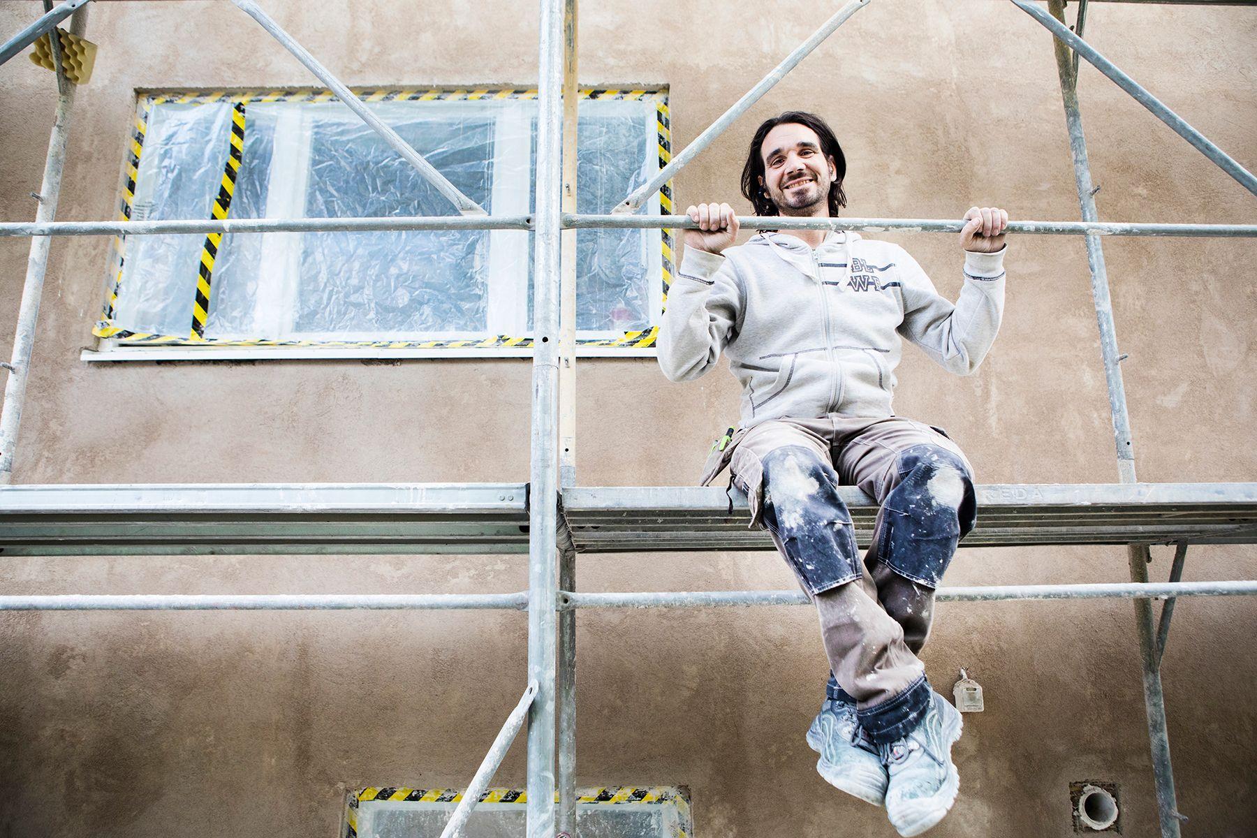 Ägaren för Grasso mur och puts iklädd arbetskläder sitter i byggställning och ler