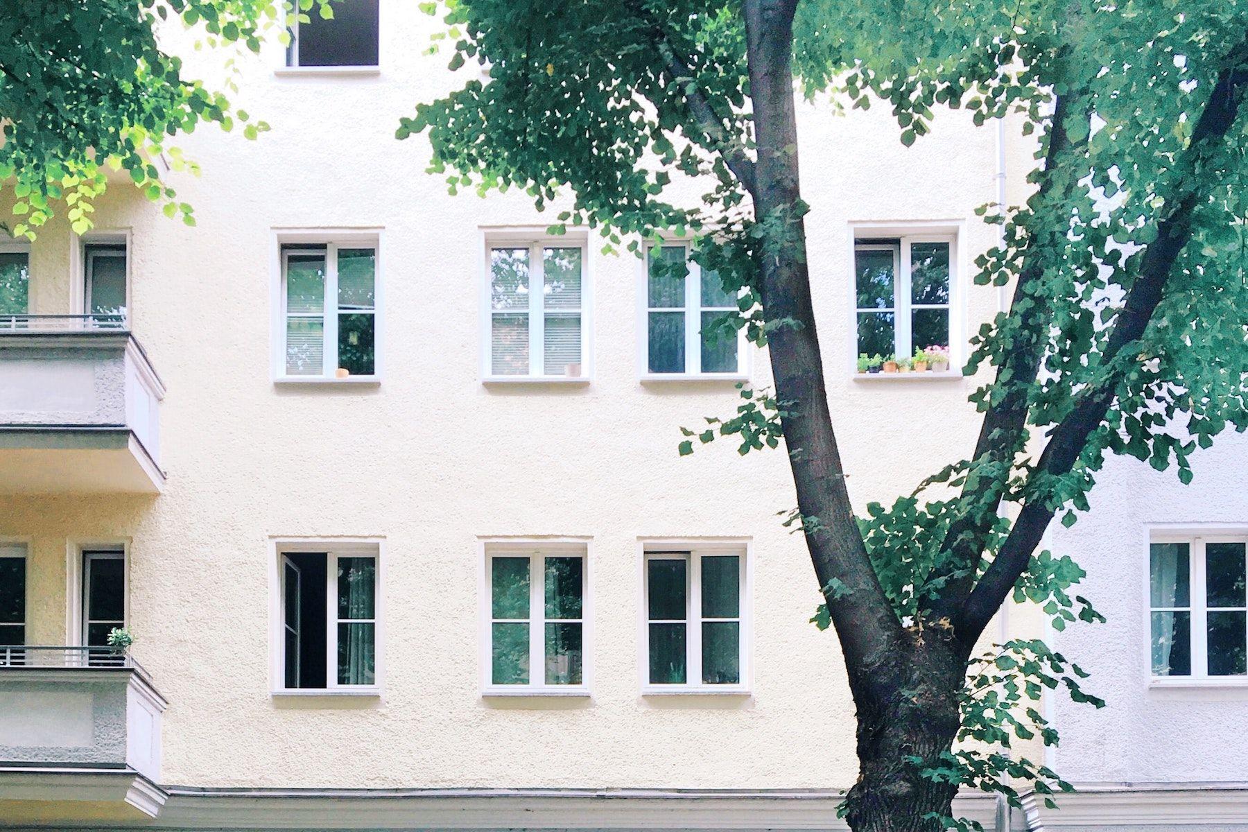 Ljust lägenhetshus med många fönster, två balkonger och grönskande träd