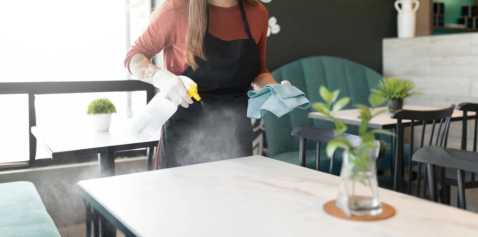 Städerska med städhandskar sprutar rengörsingsmedel på restaurangbord inför restaurangstädning