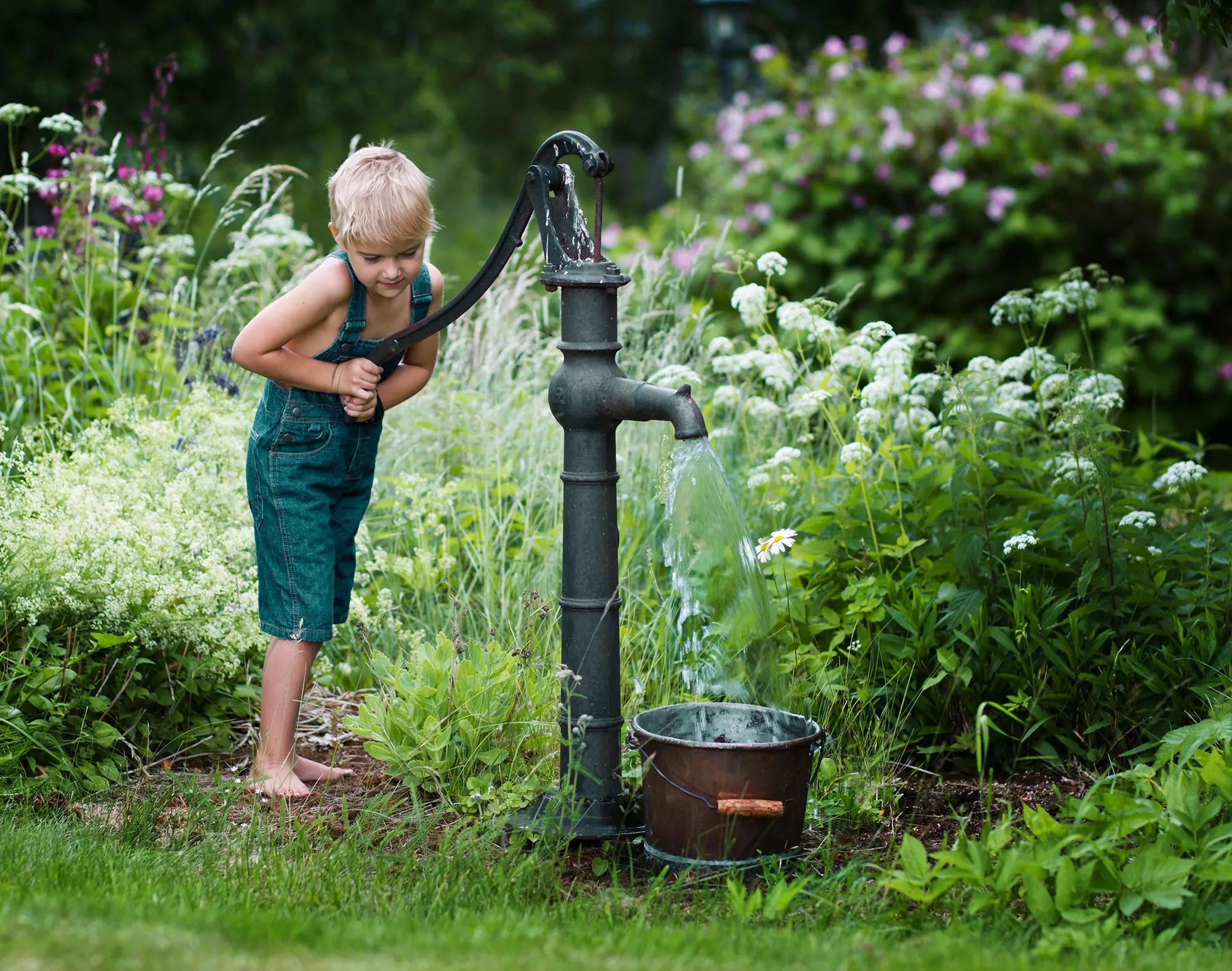 Pojke hämtar och pumpar vatten från en gammal vattenpump i idyllisk trädgård