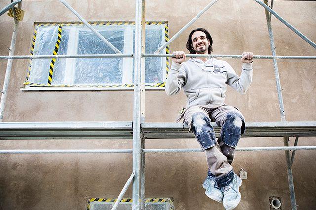 Ägaren för Grasso mur och puts iklädd arbetskläder sitter i byggställning och ler