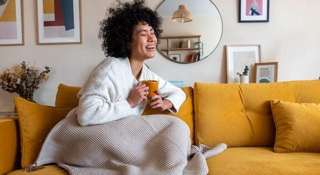 En kvinna som sitter nedbäddad under en filt i en senapsgul soffa. Hon skrattar och håller en mugg i handen.