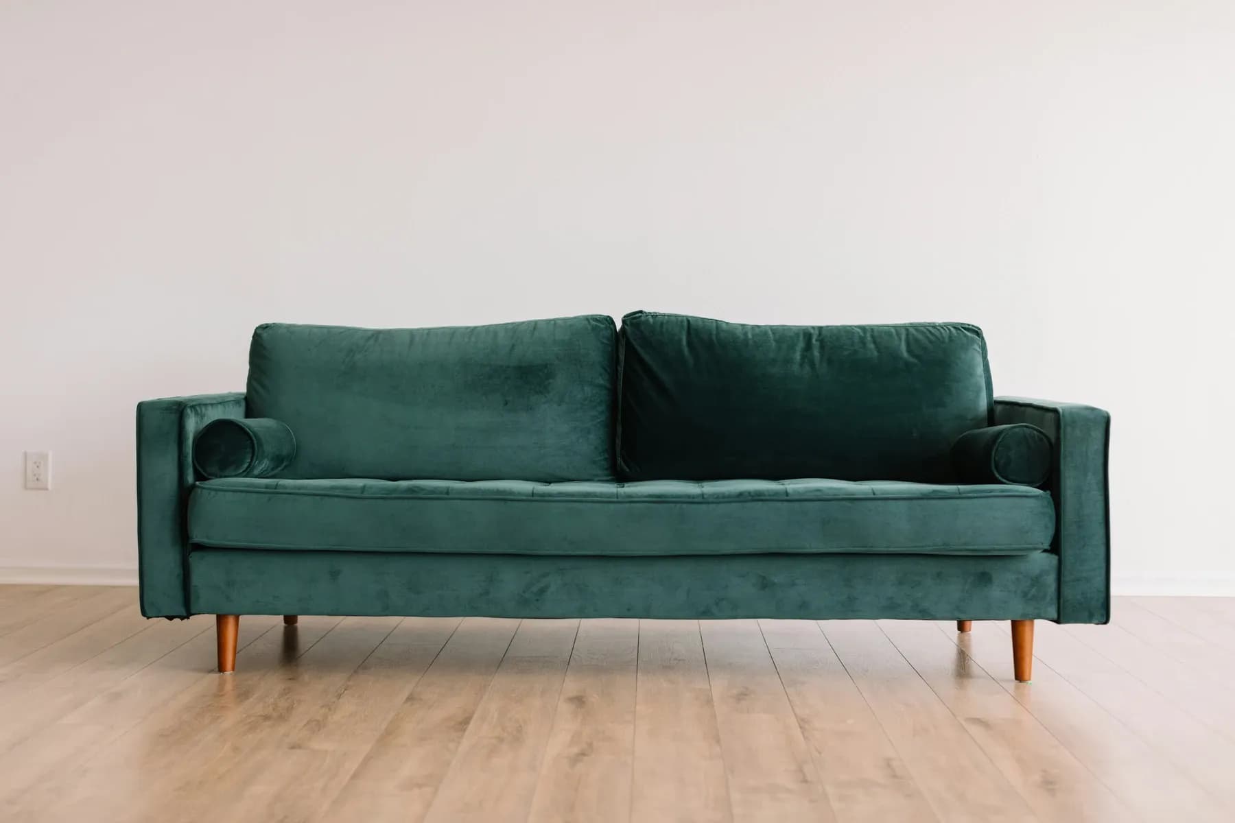 Teal velvet mid-century sofa with round bolster pillows and wooden legs, set against a plain white wall on light hardwood flooring.