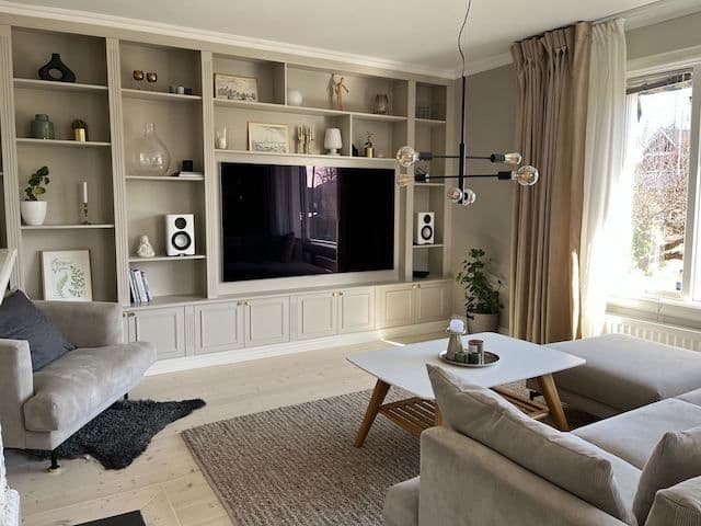 Bild från reportage om platsbyggd bokhylla. Bokhyllan är målad i en grå-beige nyans och på bilden syns soffa, soffbord och fåtölj.